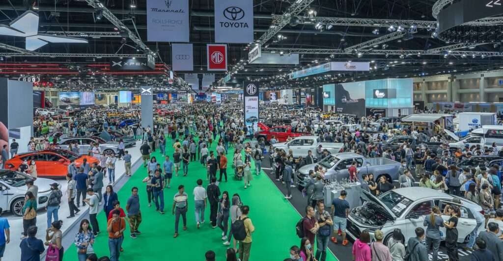 Crowded exhibition hall at the Bangkok International Motor Show with numerous automotive displays, brand banners including Maserati, Rolls-Royce, Toyota, Audi, and MG hanging from the ceiling, and attendees walking along a green carpeted central aisle.