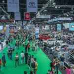 Crowded exhibition hall at the Bangkok International Motor Show with numerous automotive displays, brand banners including Maserati, Rolls-Royce, Toyota, Audi, and MG hanging from the ceiling, and attendees walking along a green carpeted central aisle.
