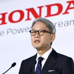 Honda CEO Toshihiro Mibe speaking at a press conference with the Honda logo and slogan "The Power of Dreams" visible in the background