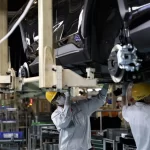 Factory workers in protective gear and yellow helmets working on vehicle assembly in a Thai automotive manufacturing plant.