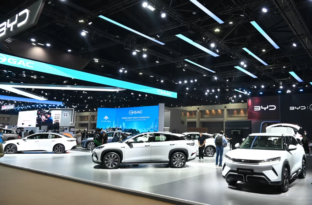 BYD electric vehicle display at an auto show featuring multiple white EVs on a sleek exhibition floor with modern blue lighting and digital screens